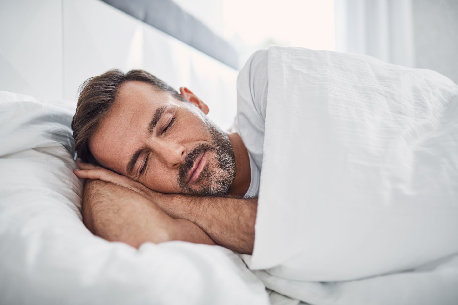 A man with a beard is sleeping in a bed with white sheets.