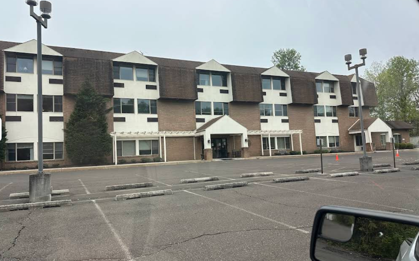 Three-story brick building with brown roof and white dormers, fronted by a large parking lot.