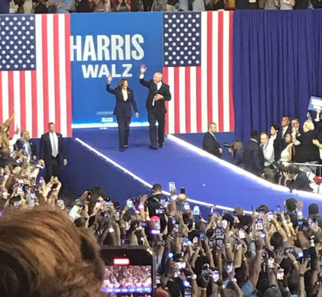 Kamala Harris and Tim Walz waving to a crowd at a rally, with American flags in the background.
