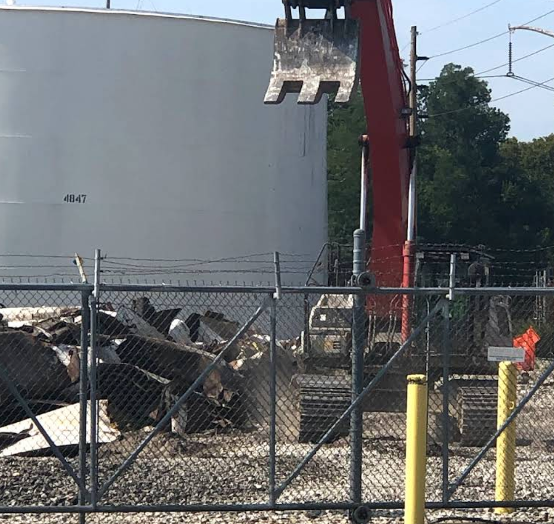 An excavator demolishes debris behind a chain-link fence, near a large white tank.
