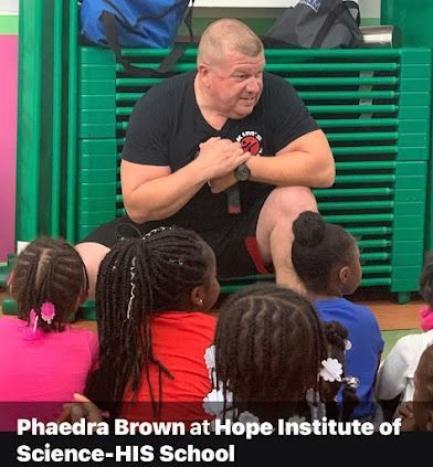 Man speaking to children at a school. He’s kneeling, wearing a black shirt. Children face the man, some wearing pink and blue.