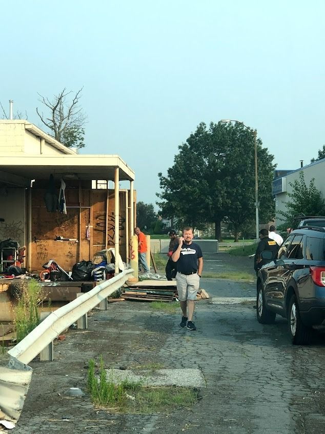 People near a damaged building with debris. One person speaks on a phone, another in an orange shirt. A car parked nearby.