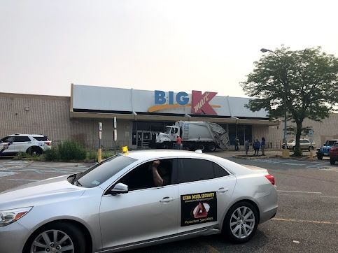 A silver car in front of a Kmart. A garbage truck is near the store entrance. People stand outside.