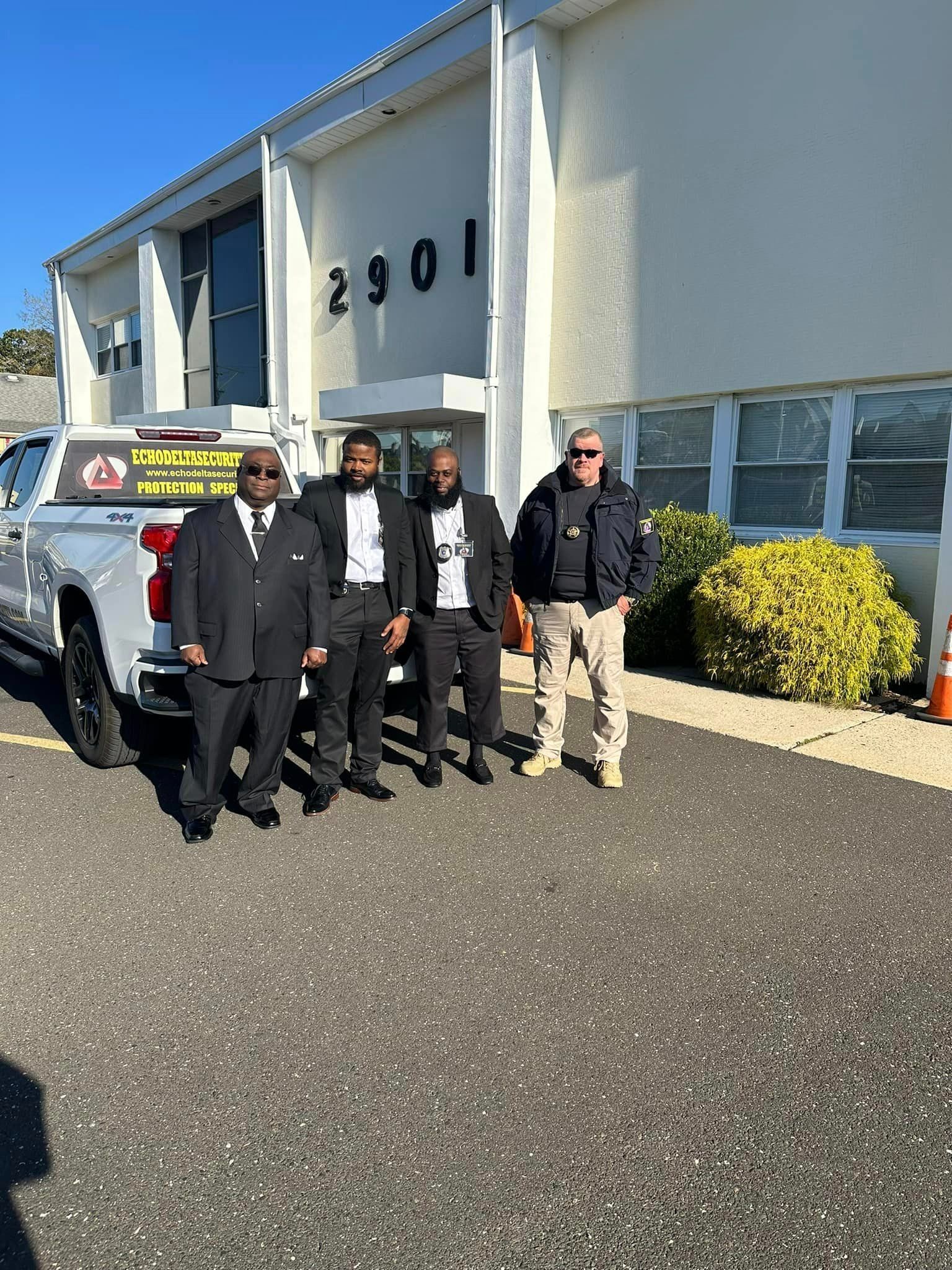Four men in suits stand in front of a building with a white truck.