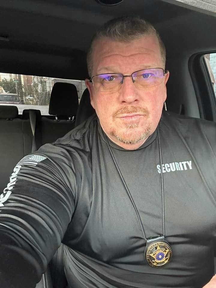Man in a security uniform wearing glasses, badge, and looking at the camera. He is inside a vehicle.