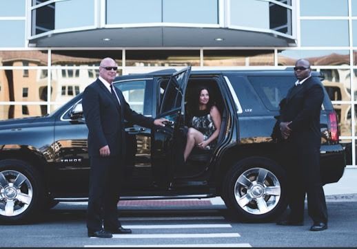 Two bodyguards assist a person entering a black SUV. Modern building in the background.