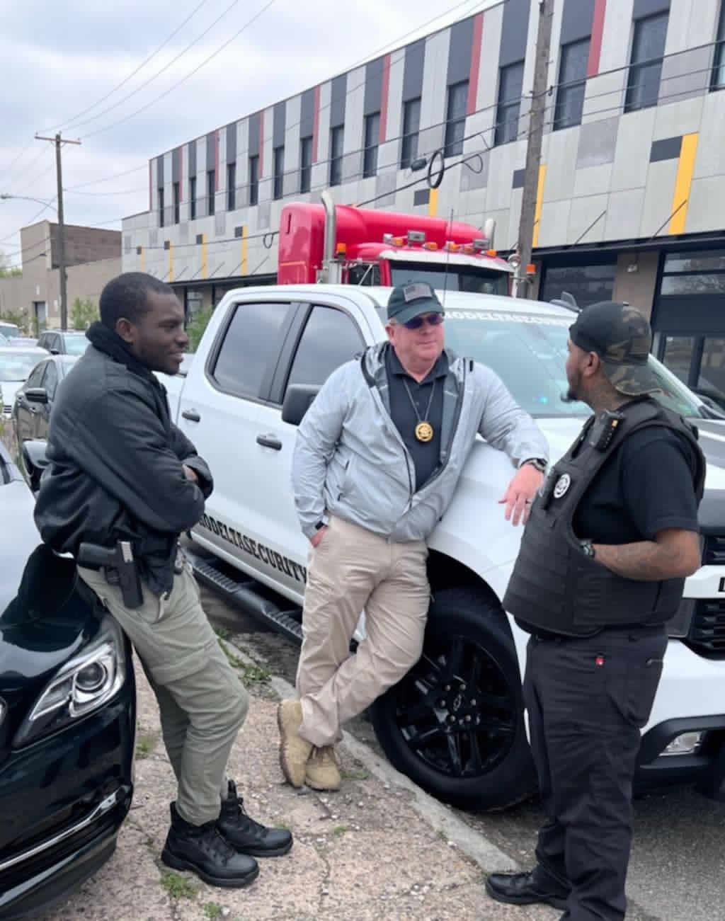 Three men standing near a white pickup truck. Two lean against vehicles; one wears a vest. Cloudy day.