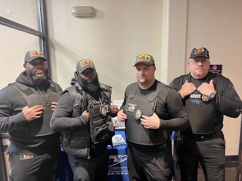 Four uniformed men wearing vests and hats, posing indoors. They are in a well-lit area with a table set up.