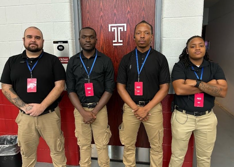 Four people in black shirts and khaki pants stand in front of a Temple University door.