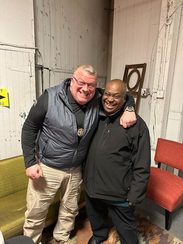 Two men smiling, arm in arm. One wears a vest over a black shirt and khaki pants, the other a black jacket. Indoors, against a wall.