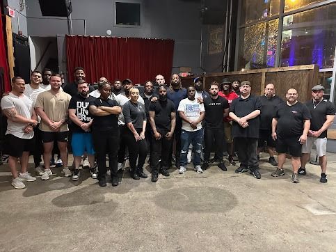 Group of people indoors, some in black shirts. Standing in front of a red curtain and a concrete floor.