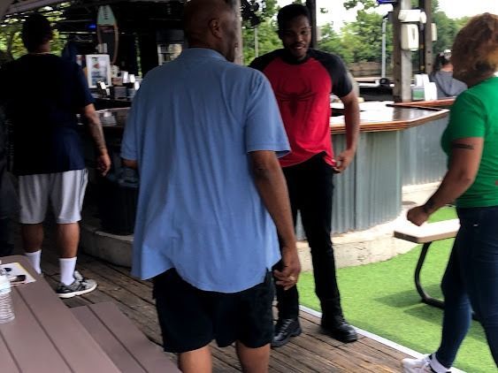 People conversing at an outdoor bar. One man in blue shirt, another in a red and black shirt. Others near a bar and tables.