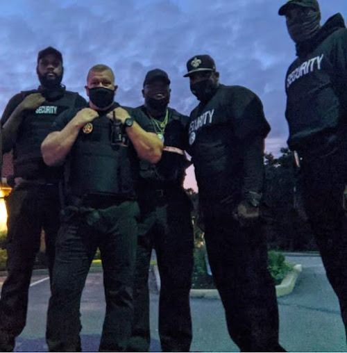 Five security guards in black attire, posed outdoors, under a dusky sky, some wearing masks.