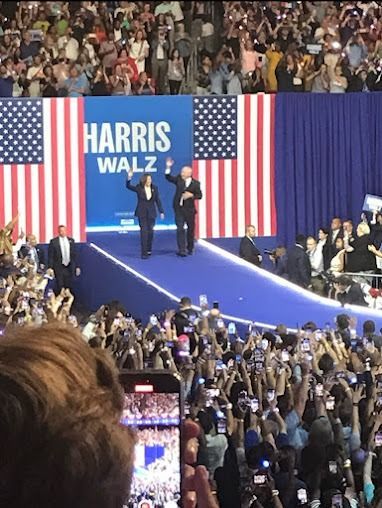Harris and Walz on stage waving to a large crowd, banner says 