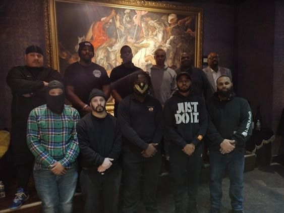 Group of people posing in front of a painting inside a building. Some have covered faces.