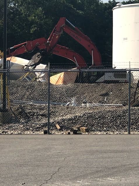 Red excavator demolishing a building behind a chain-link fence, with gravel and debris visible.