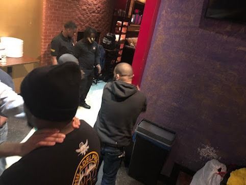 People interacting inside a dimly lit interior. Several in dark uniforms, one wearing a black beanie, near a purple wall.