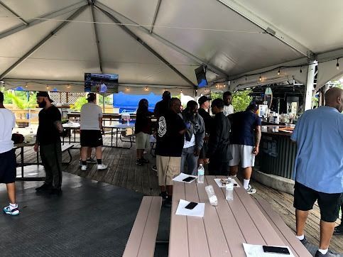 Group of people under a tented patio, some standing at the bar, others at picnic tables, with a TV on the wall.
