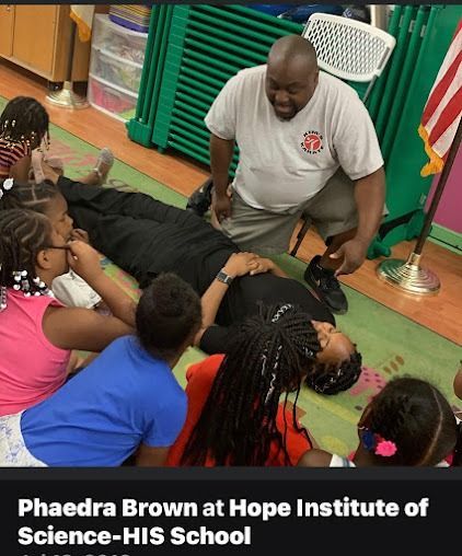 Man kneels by person on floor surrounded by children at school.