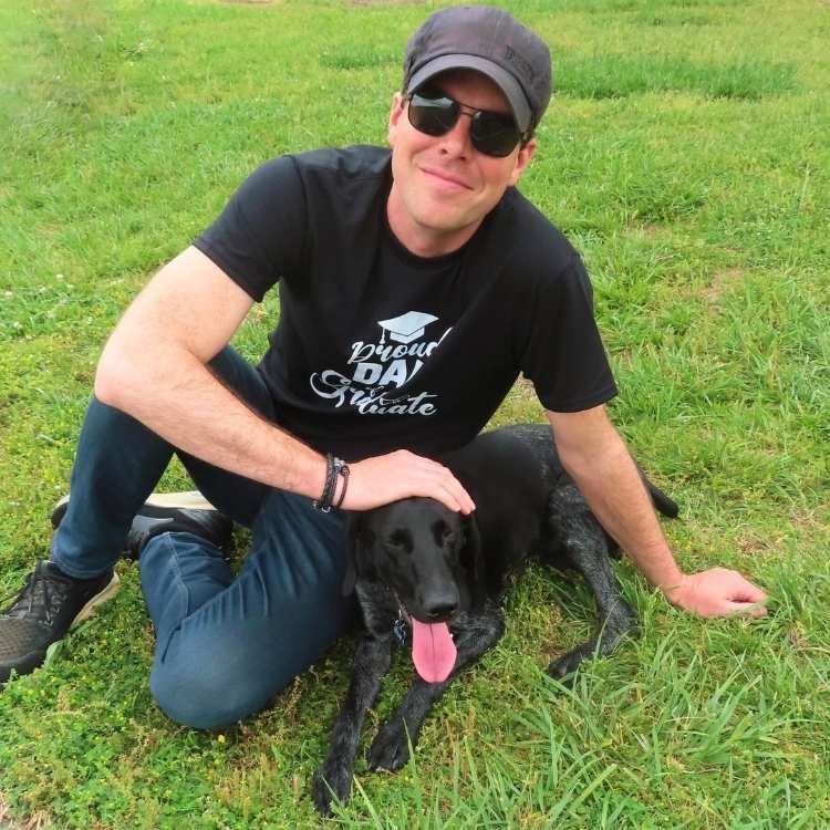 A man wearing a black shirt that says proud dad kneeling next to a black dog