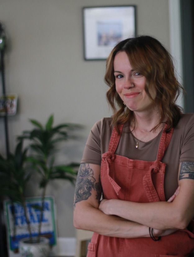 A woman in an apron is standing in a living room with her arms crossed.