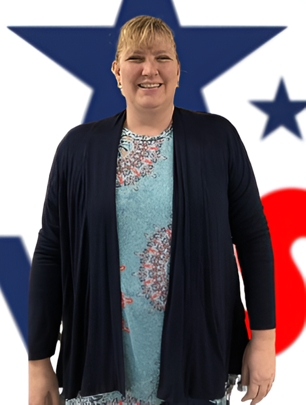 A woman wearing a blue shirt and a black cardigan is standing in front of an american flag.