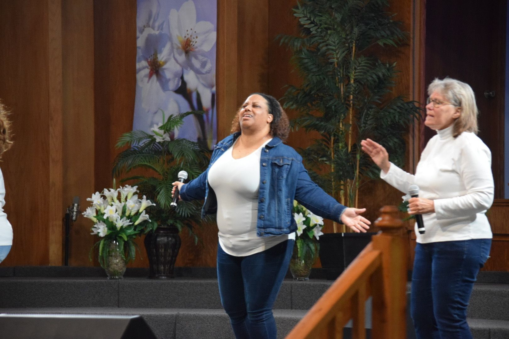 Two women singing on a stage. One with arms open, other gesturing, both holding microphones.