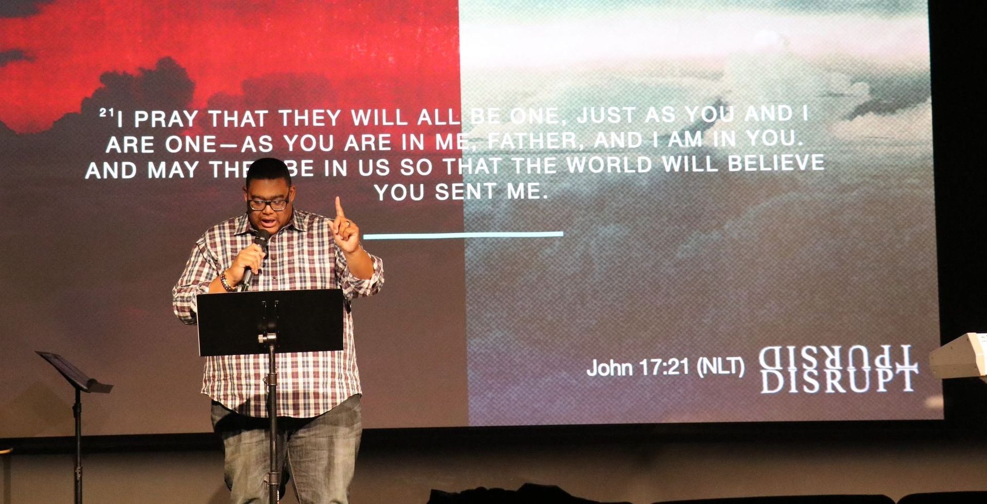 Man speaking at a podium with Bible verse on a screen.