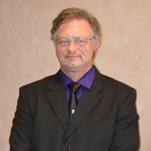 Man wearing glasses, black suit, purple shirt, and tie; looking at the camera.