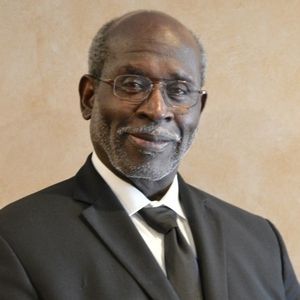 Man wearing glasses, black suit, and tie, smiling, indoors.