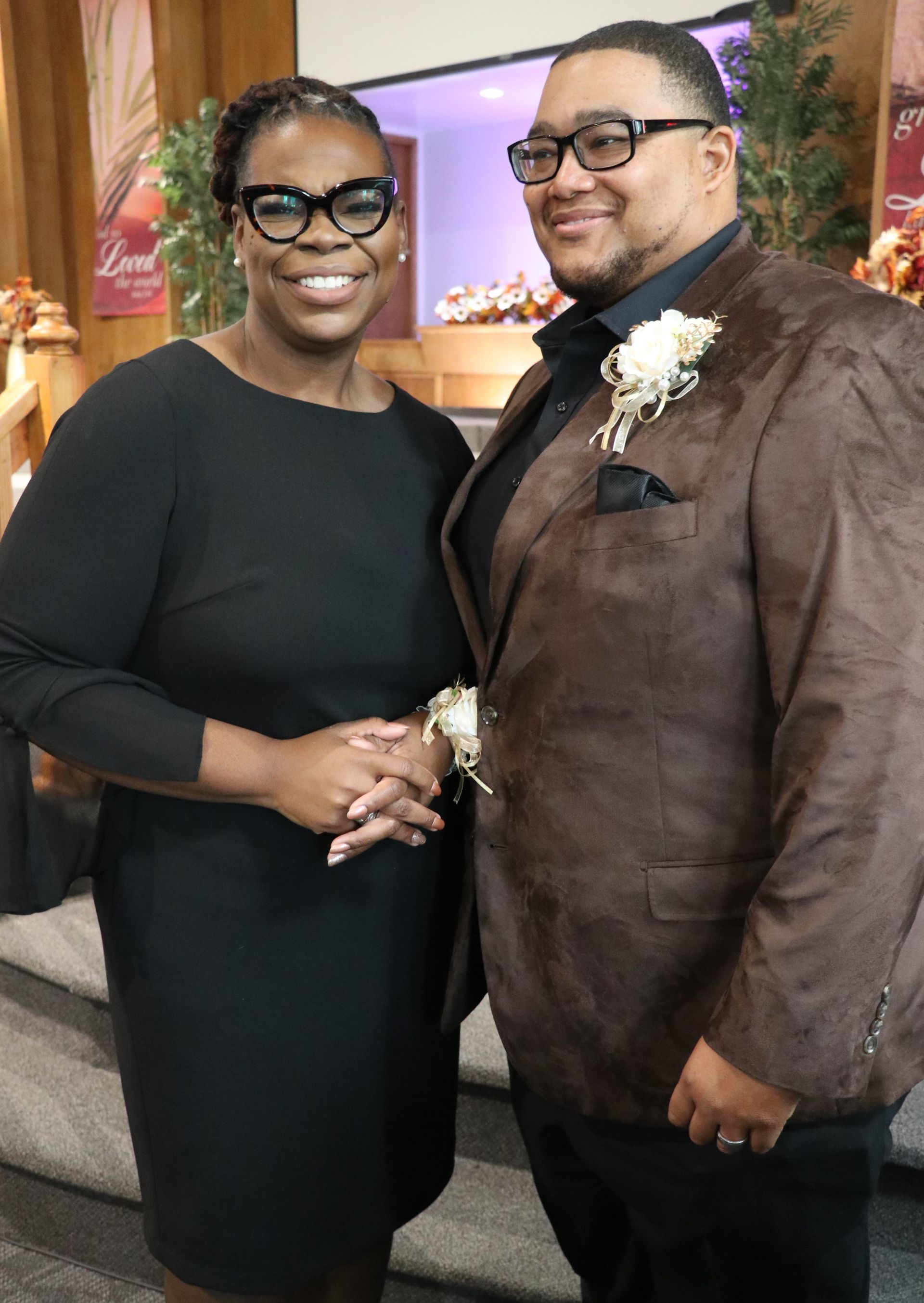 Woman in black dress and man in brown jacket pose together indoors.