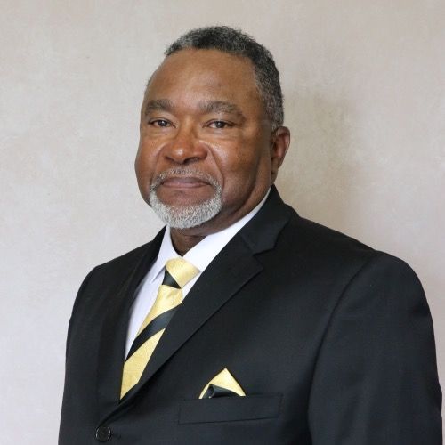 Man in black suit with gold tie and pocket square, posing in front of a neutral backdrop.