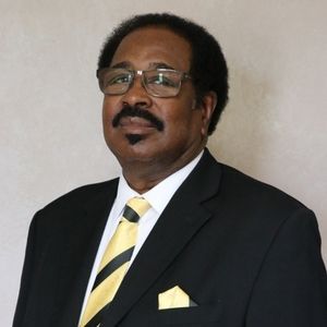 Man in black suit, glasses, and yellow tie, smiling.