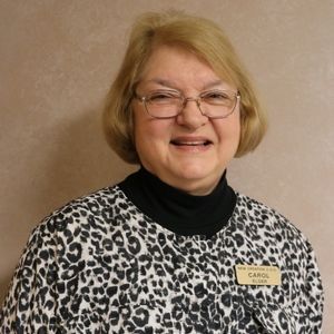 Woman wearing glasses, smiling, with blonde hair, black turtleneck, and black/white patterned top. Name tag: Carol.