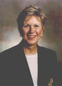 Woman with short blonde hair smiles, wearing a dark blazer over a white blouse, studio background.