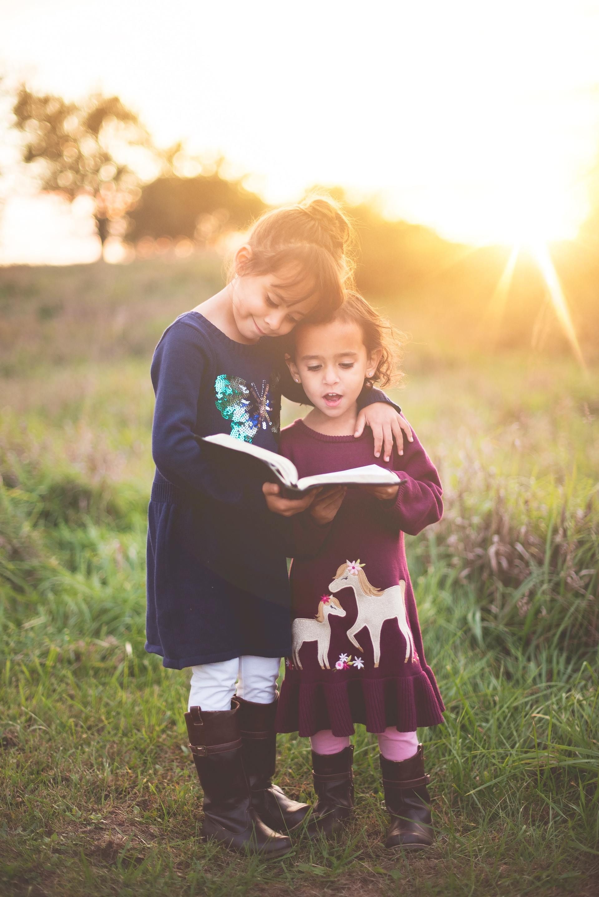 Two young girls reading a book together in a field at sunset; one has an arm around the other.