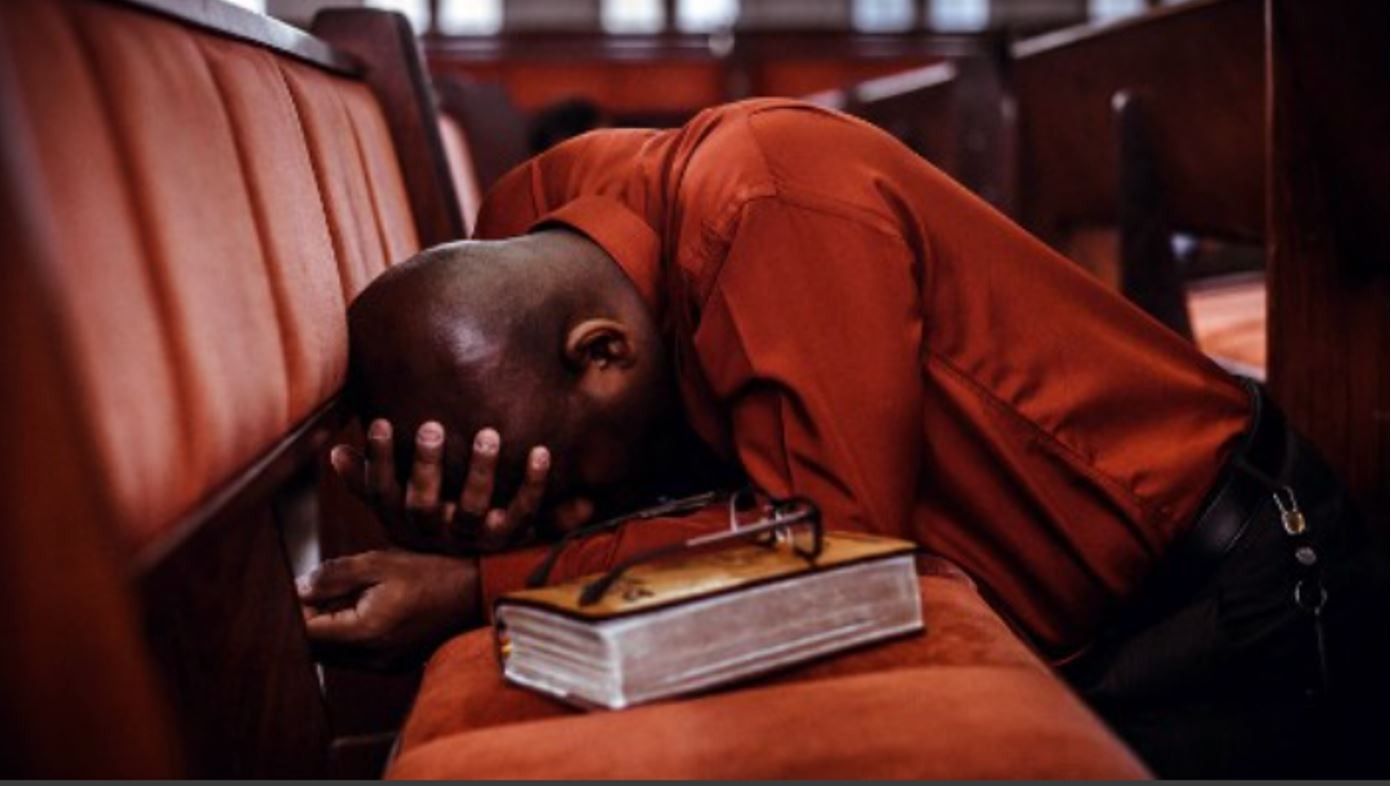 Man in red shirt kneels in church pew, head bowed on a bible, hands covering face.