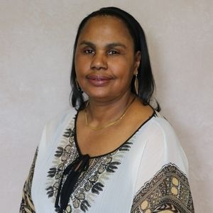 Woman wearing a white patterned top, smiling, gold necklace and earrings.