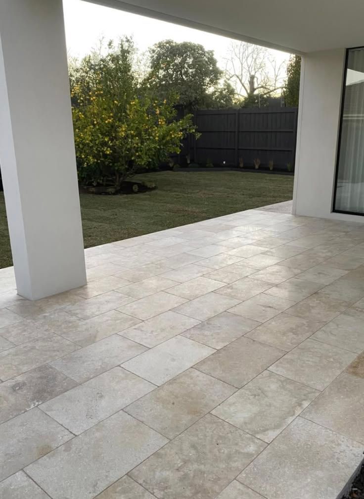 Covered patio with beige tile flooring, white columns, and a grassy backyard with trees and a dark fence.