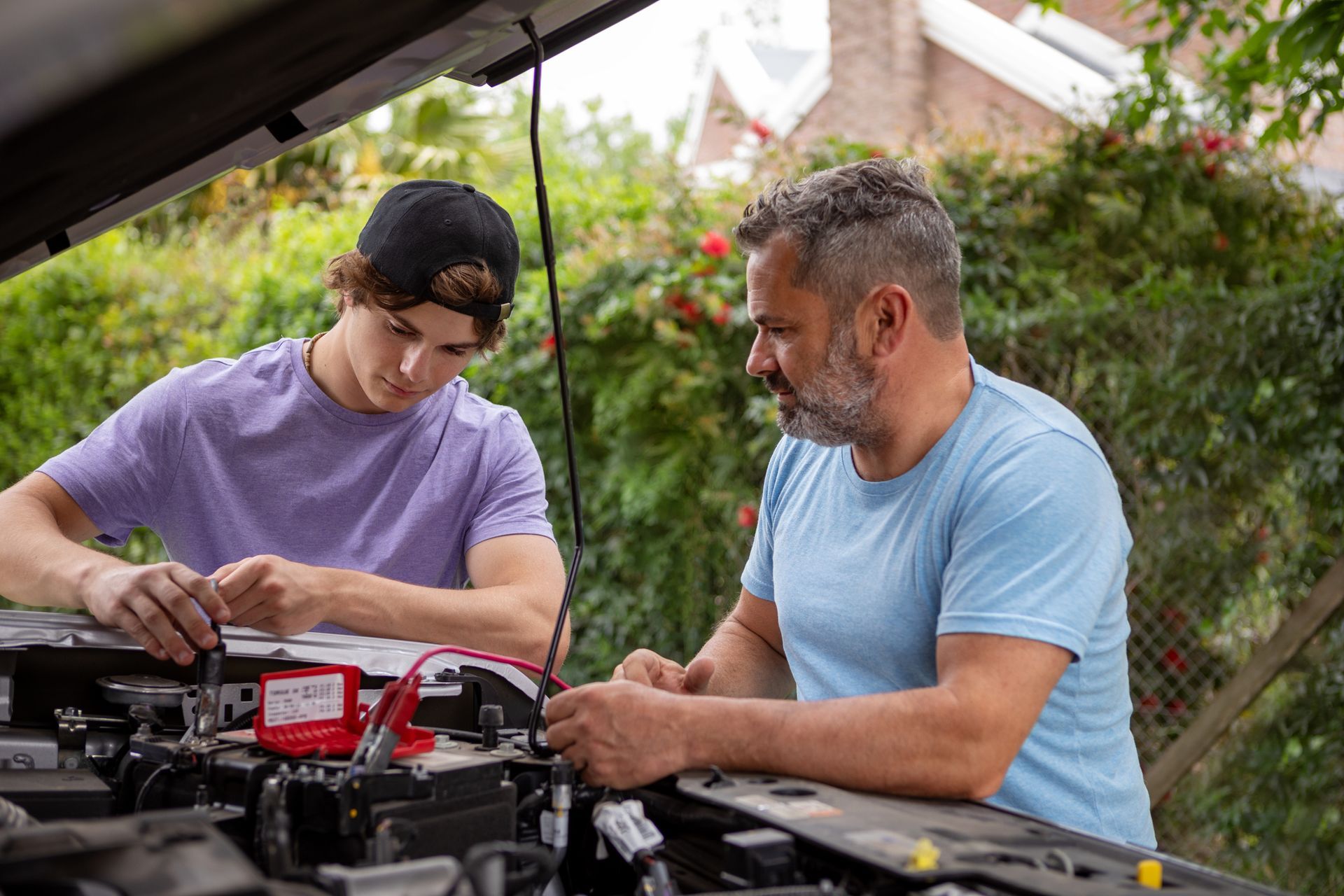 Man and teen working on a car engine, outdoors. They are holding tools, assessing the mechanics.