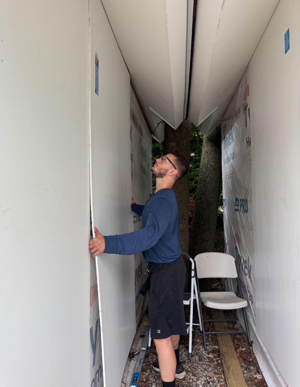 Man installing drywall white panels around a tree, standing in narrow outdoor space, looking up.