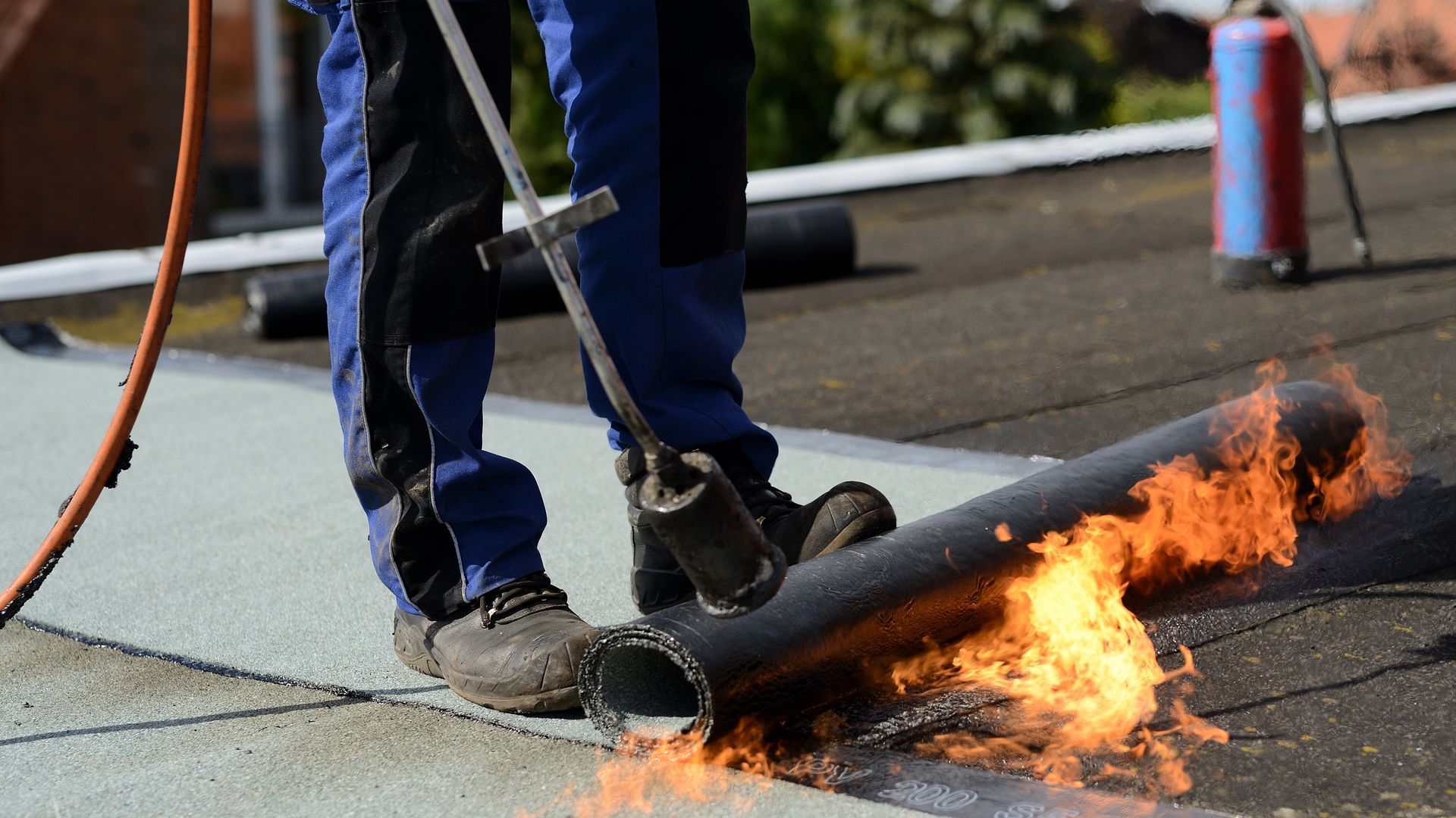 A man is using a torch to burn a roll of roofing material on a roof.
