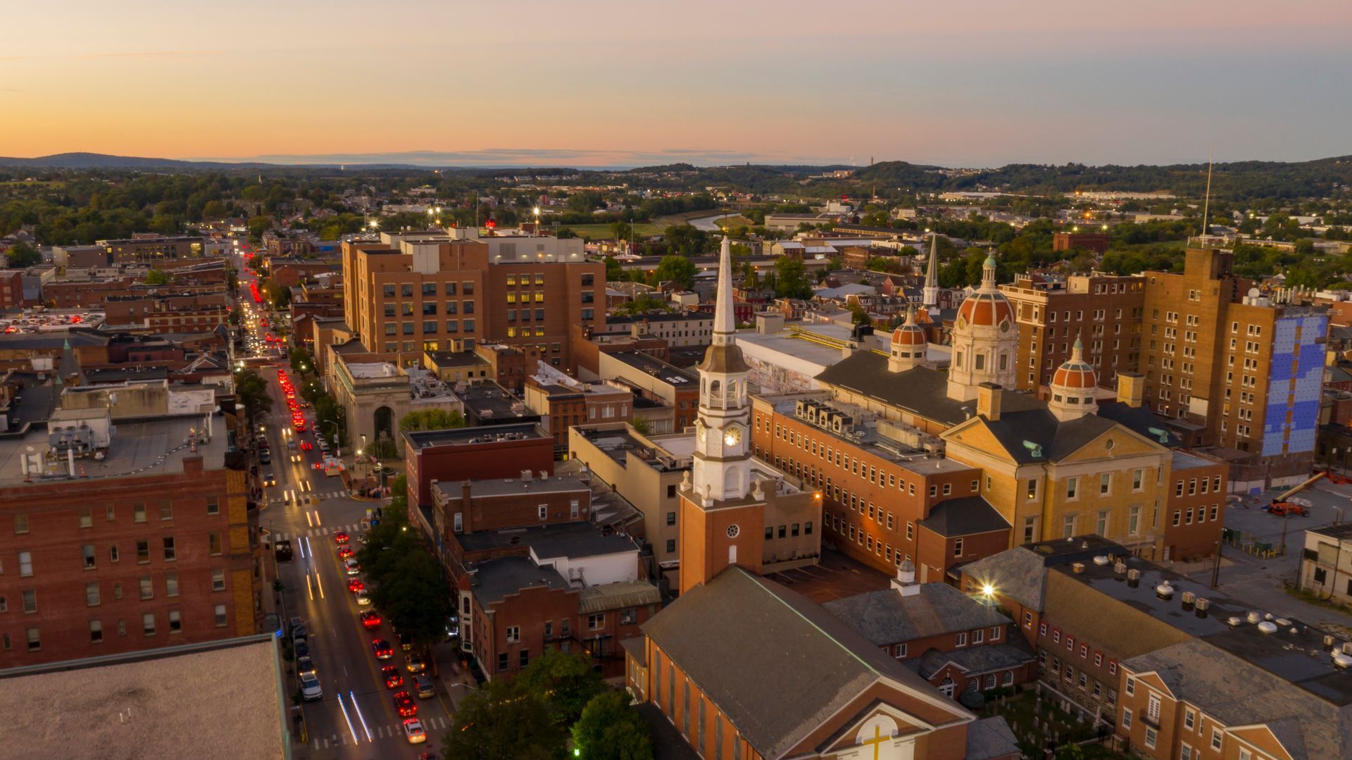 Aerial view of a city at sunset with brick buildings, a church steeple, and traffic on a main street.