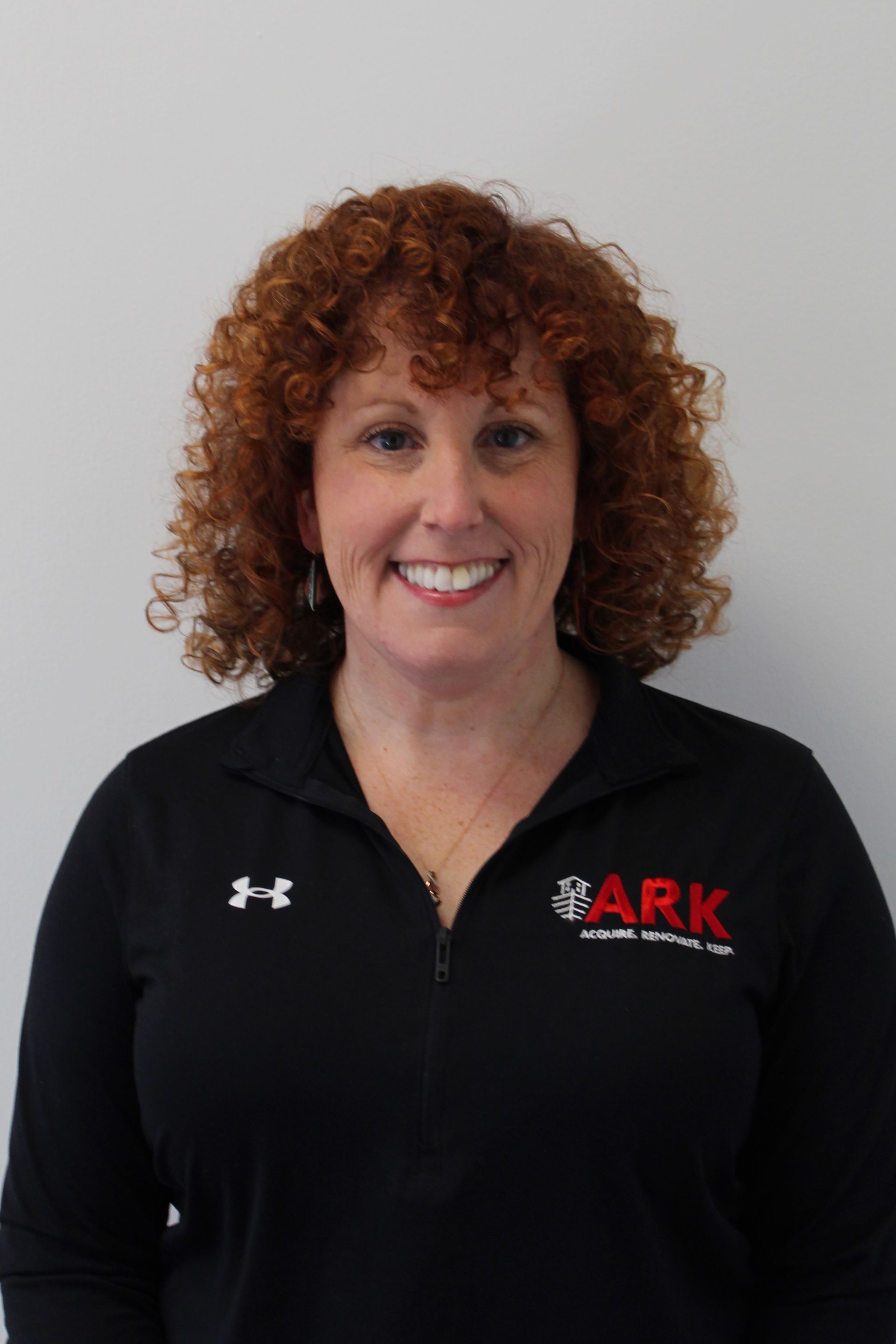 Woman with red curly hair wearing a black shirt smiling, with the ARK logo.