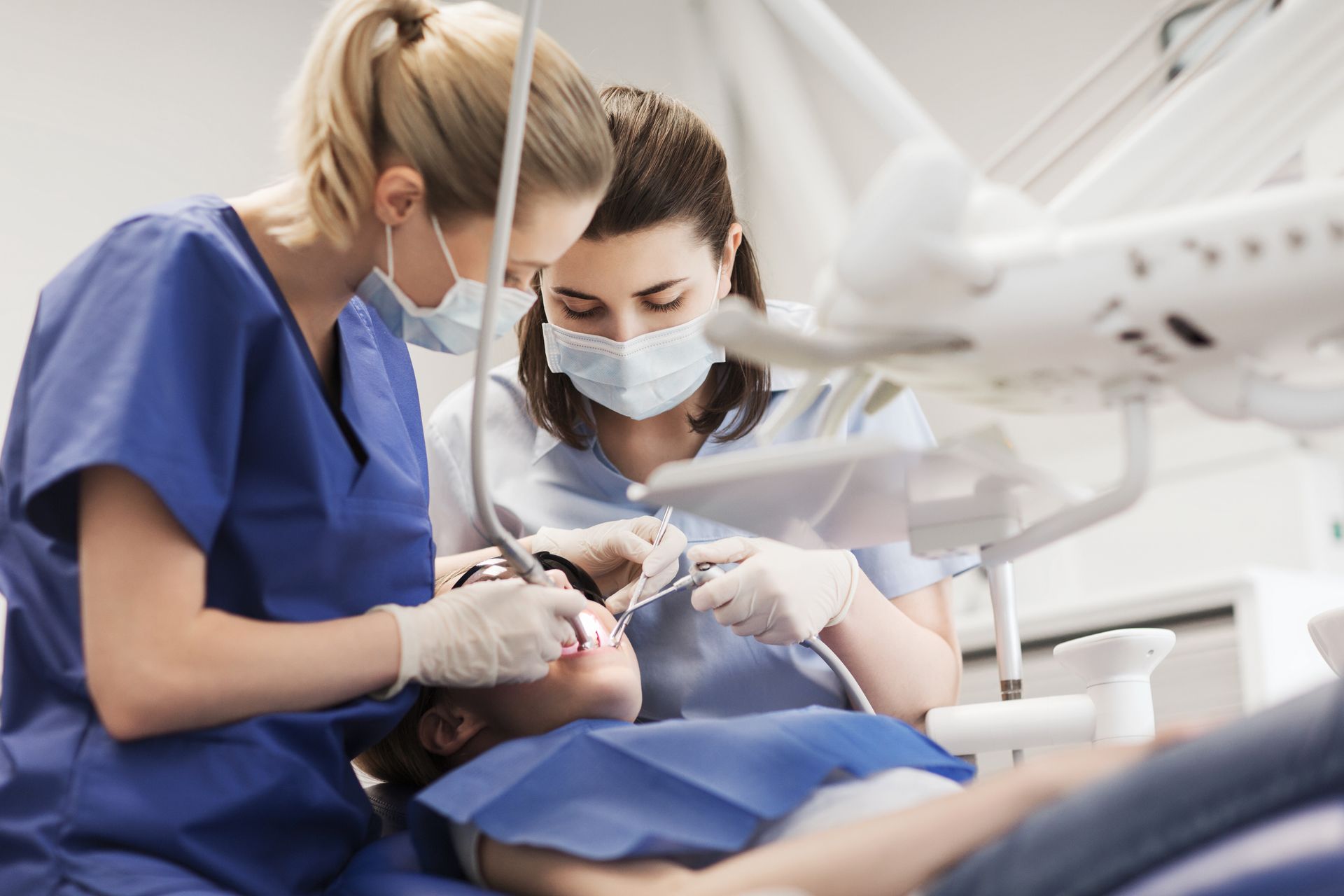 Dentists in blue scrubs treating a patient in a dental clinic, with one using tools and the other assisting.