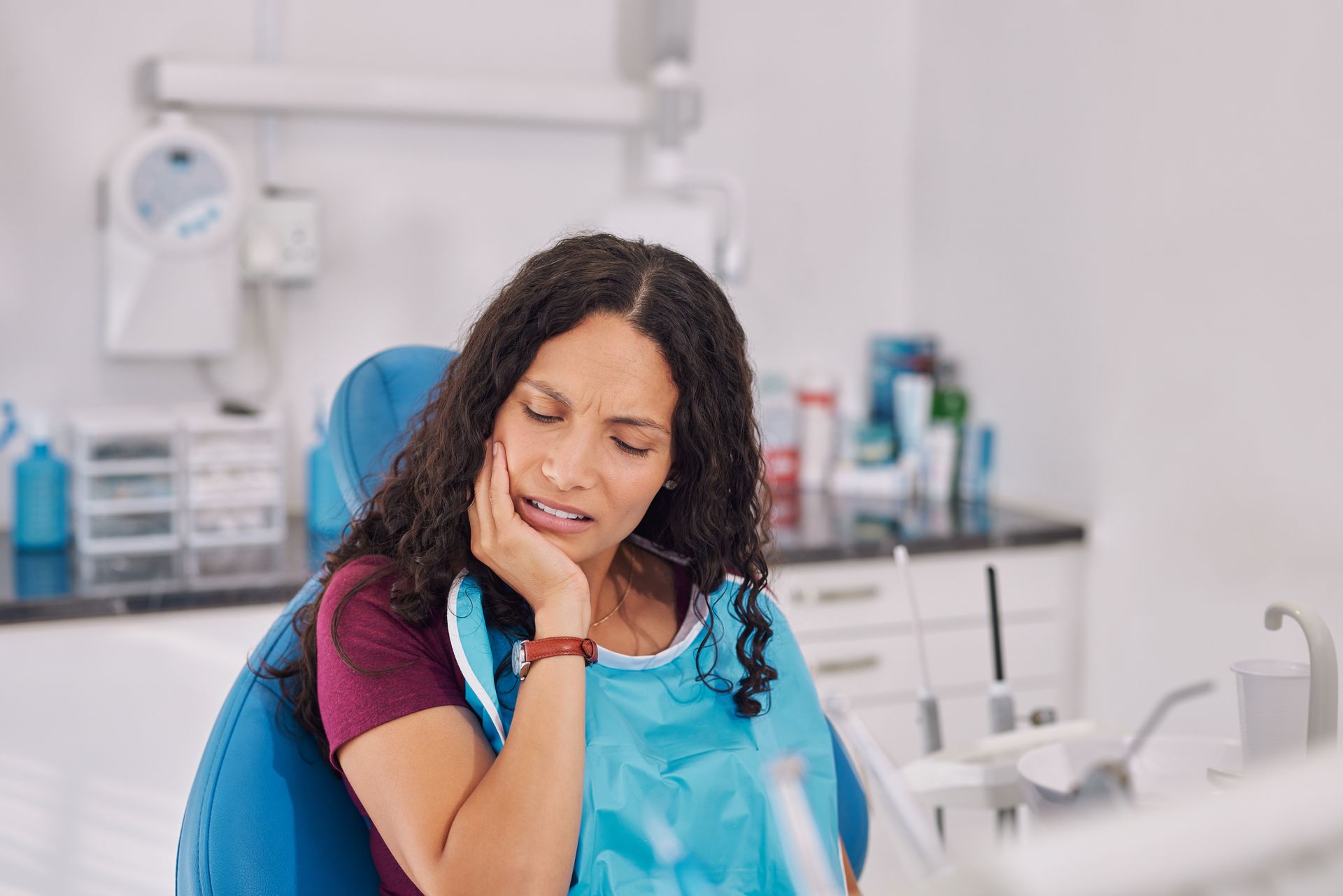 Woman in a dental chair with blue bib, holding her cheek in a bright clinic