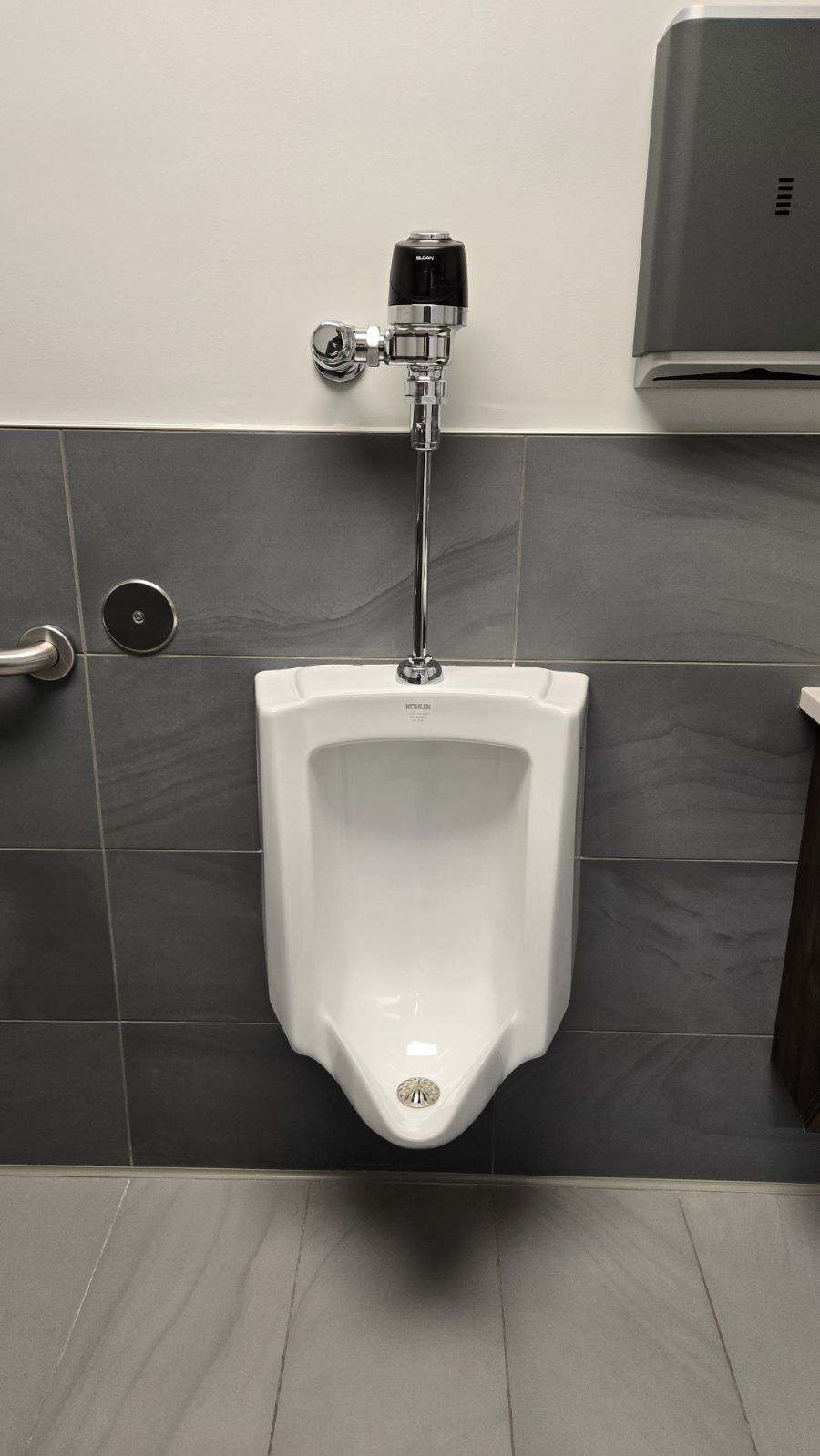 White urinal on gray tiled wall with chrome fixtures.