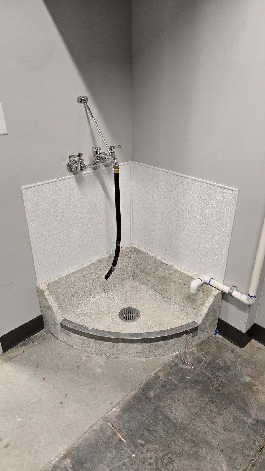 Corner pet wash station with hose and drain, surrounded by white speckled tile. Gray walls and flooring.