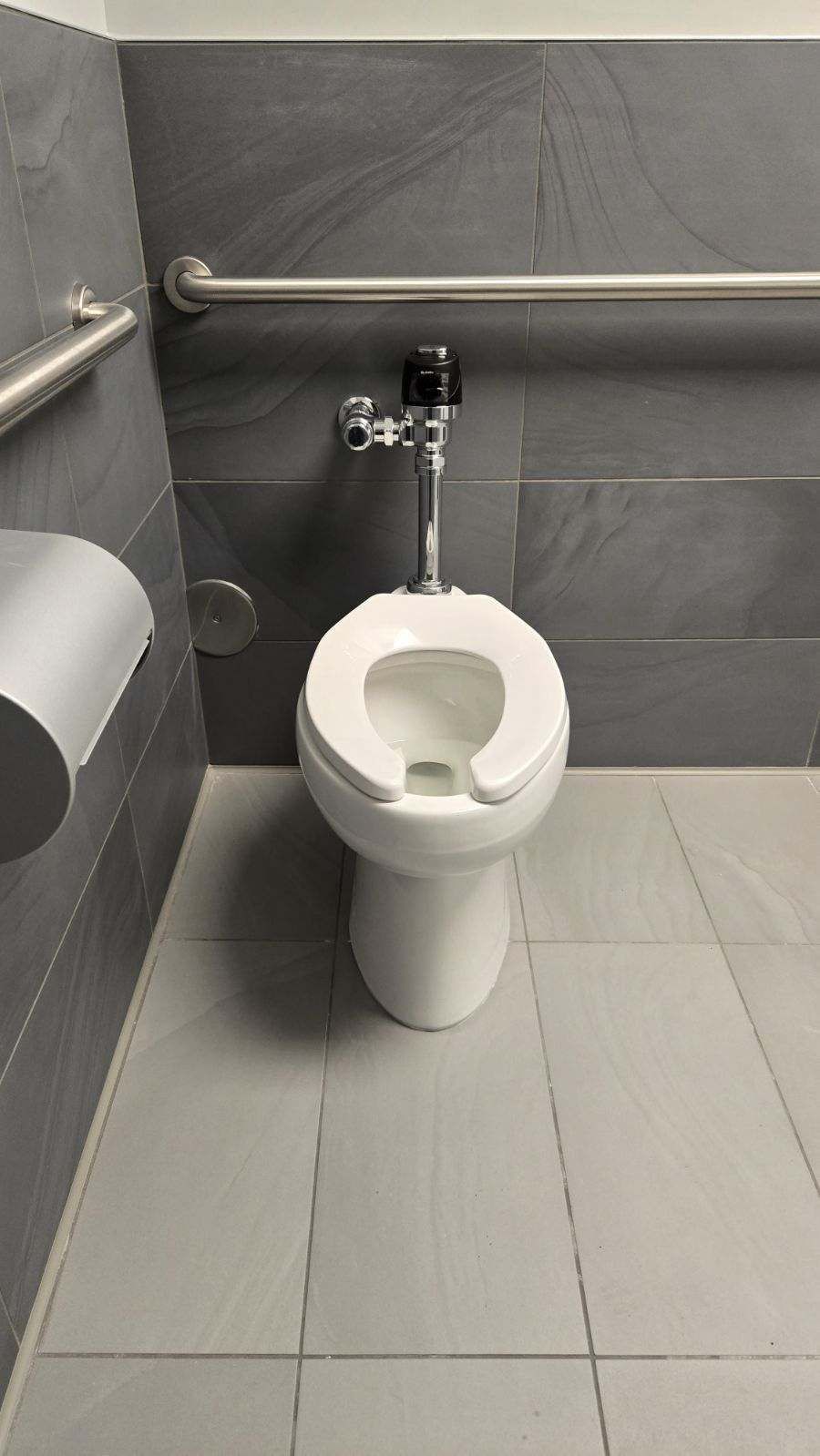 Toilet in a public restroom with gray tiled walls and a grab bar.