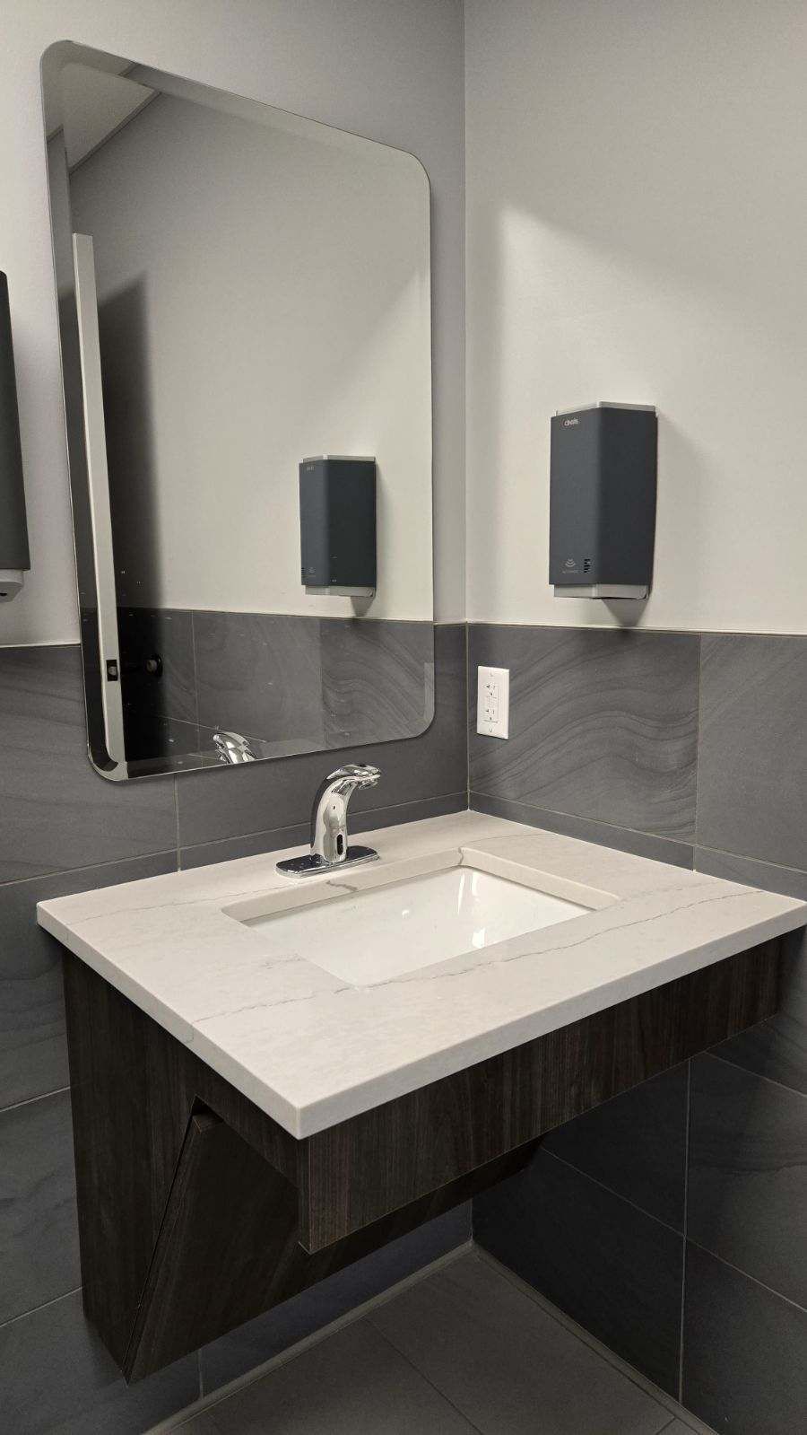 A wall-mounted sink with a large mirror, two soap dispensers, and gray tiled walls in a bathroom.